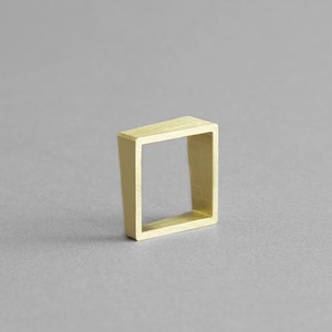 Op de afbeelding: Een gouden ring met een vierkant ontwerp. De ring is gemaakt van een dikke metalen band met een hol midden.