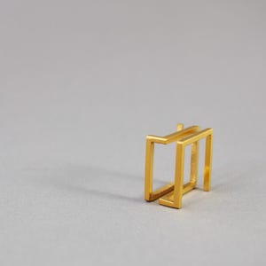 May include: A gold-toned ear cuff featuring a double-layered rectangular frame design. The piece consists of two parallel, open-ended square bars connected at the top, creating a geometric structure with a smooth, metallic finish and sharp edges.