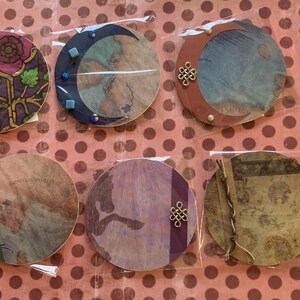 May include: A collection of ten round decorative items, each with unique designs and embellishments. The items feature various colors, patterns, and textures, including wood grain, floral motifs, and celestial themes. Some are adorned with metal accents.