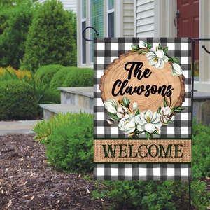 May include: A black and white checkered garden flag with a woodgrain circle that says "The Clawson's" surrounded by white flowers and a burlap banner that says "WELCOME".