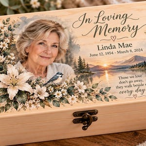 May include: A rectangular light-colored wooden keepsake box with a metal latch. The lid features a printed portrait of a person, a landscape with a lake and mountains, white floral accents, and the text In Loving Memory Linda Mae June 12, 1954 - March 8, 2024, and Those we love don't go away, they walk beside us every day.