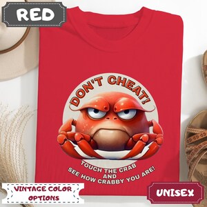 May include: Red unisex t-shirt featuring a cartoon crab with the text "DON'T CHEAT! TOUCH THE CRAB AND SEE HOW CRABBY YOU ARE!". The shirt has "VINTAGE COLOR OPTIONS" and "UNISEX" labels.