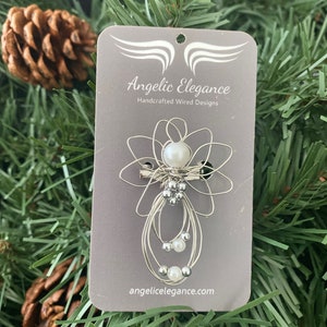 May include: A silver-toned wire angel pin with a pearl head and body, and small silver beads. The pin is attached to a gray card with the text "Angelic Elegance Handcrafted Wired Designs" and the website address angelicelegance.com.