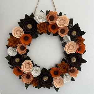 Puede incluir: Una corona de flores de fieltro hecha a mano con flores marrones, naranjas, melocot&oacute;n y blancas. La corona est&aacute; acentuada con hojas verdes oscuras y peque&ntilde;os elementos decorativos, suspendida por una cuerda natural.
