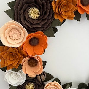 Puede incluir: Una corona de flores de fieltro hecha a mano con flores en tonos marr&oacute;n, naranja, crema y blanco. Las flores tienen hojas de fieltro verde y centros de cuentas doradas, creando un dise&ntilde;o r&uacute;stico y elegante.