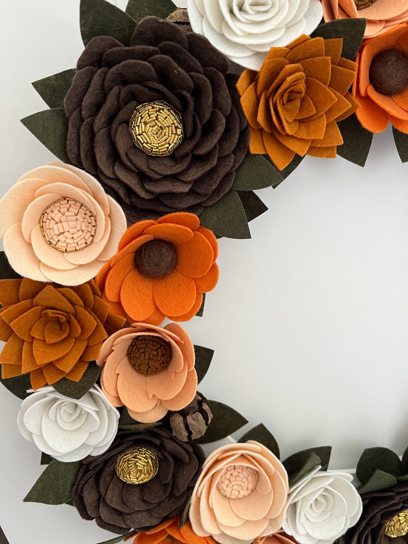 Puede incluir: Una corona de flores de fieltro hecha a mano con flores en tonos marr&oacute;n, naranja, crema y blanco. Las flores tienen hojas de fieltro verde y centros de cuentas doradas, creando un dise&ntilde;o r&uacute;stico y elegante.