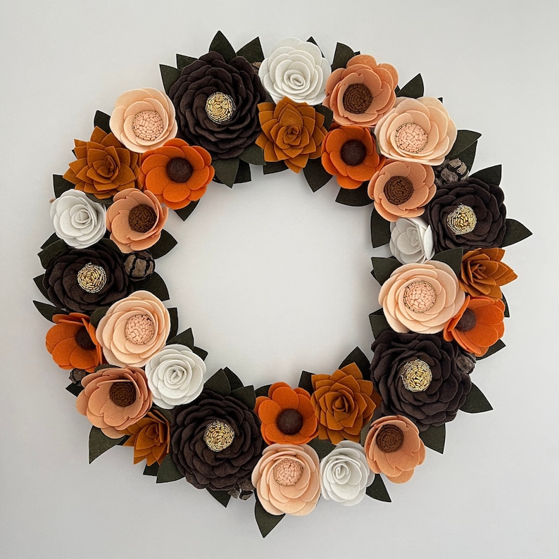 Puede incluir: Una corona de flores de fieltro hecha a mano con flores en tonos naranja, marr&oacute;n, melocot&oacute;n y blanco. La corona est&aacute; adornada con hojas de color verde oscuro y peque&ntilde;os acentos de pi&ntilde;as, creando una est&eacute;tica oto&ntilde;al c&aacute;lida.