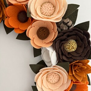 Puede incluir: Primer plano de una corona de flores de fieltro hecha a mano. La corona presenta varias flores de fieltro en tonos marr&oacute;n, naranja y crema, con hojas de color verde oscuro. Algunos centros de flores tienen detalles de cuentas doradas. La corona es un art&iacute;culo decorativo.