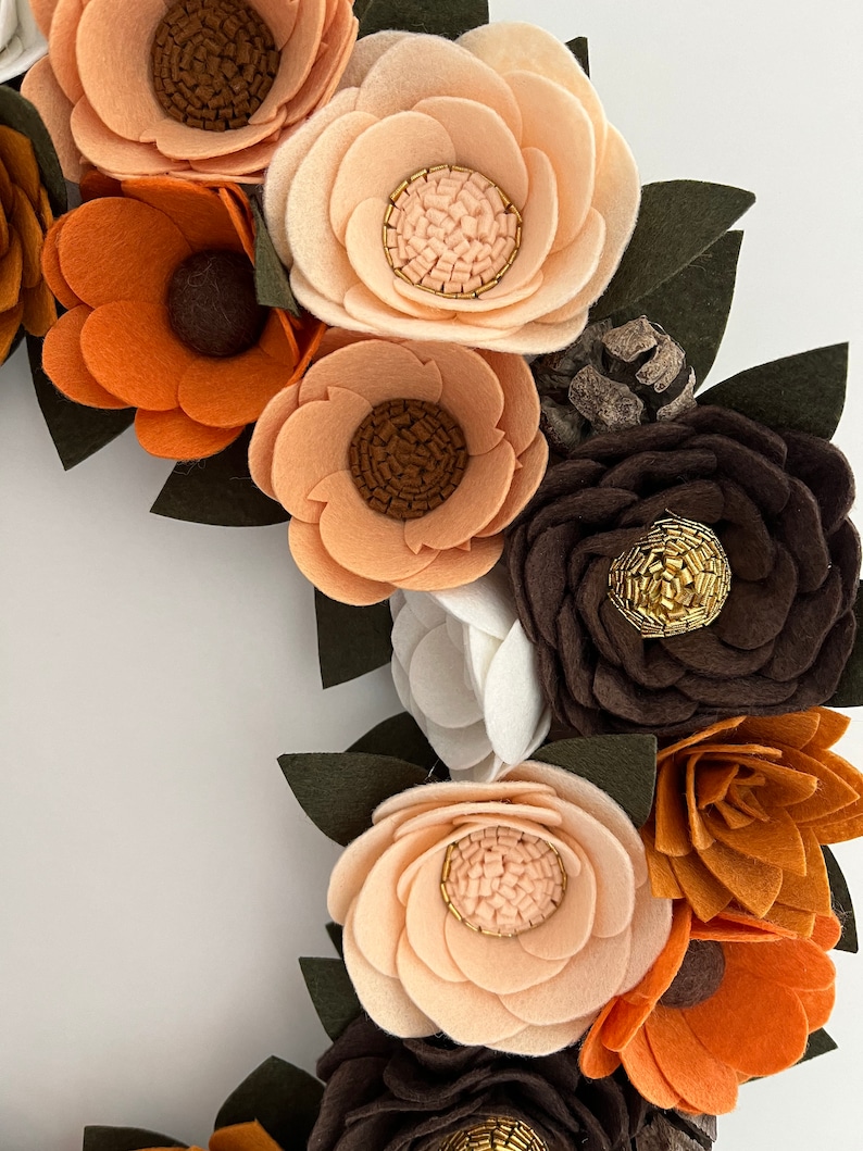 Puede incluir: Primer plano de una corona de flores de fieltro hecha a mano. La corona presenta varias flores de fieltro en tonos marr&oacute;n, naranja y crema, con hojas de color verde oscuro. Algunos centros de flores tienen detalles de cuentas doradas. La corona es un art&iacute;culo decorativo.