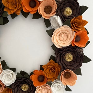 Puede incluir: Una corona de flores de fieltro hecha a mano con flores en tonos marr&oacute;n, naranja y crema, acentuada con hojas de color verde oscuro. La corona circular es ideal para la decoraci&oacute;n de temporada.