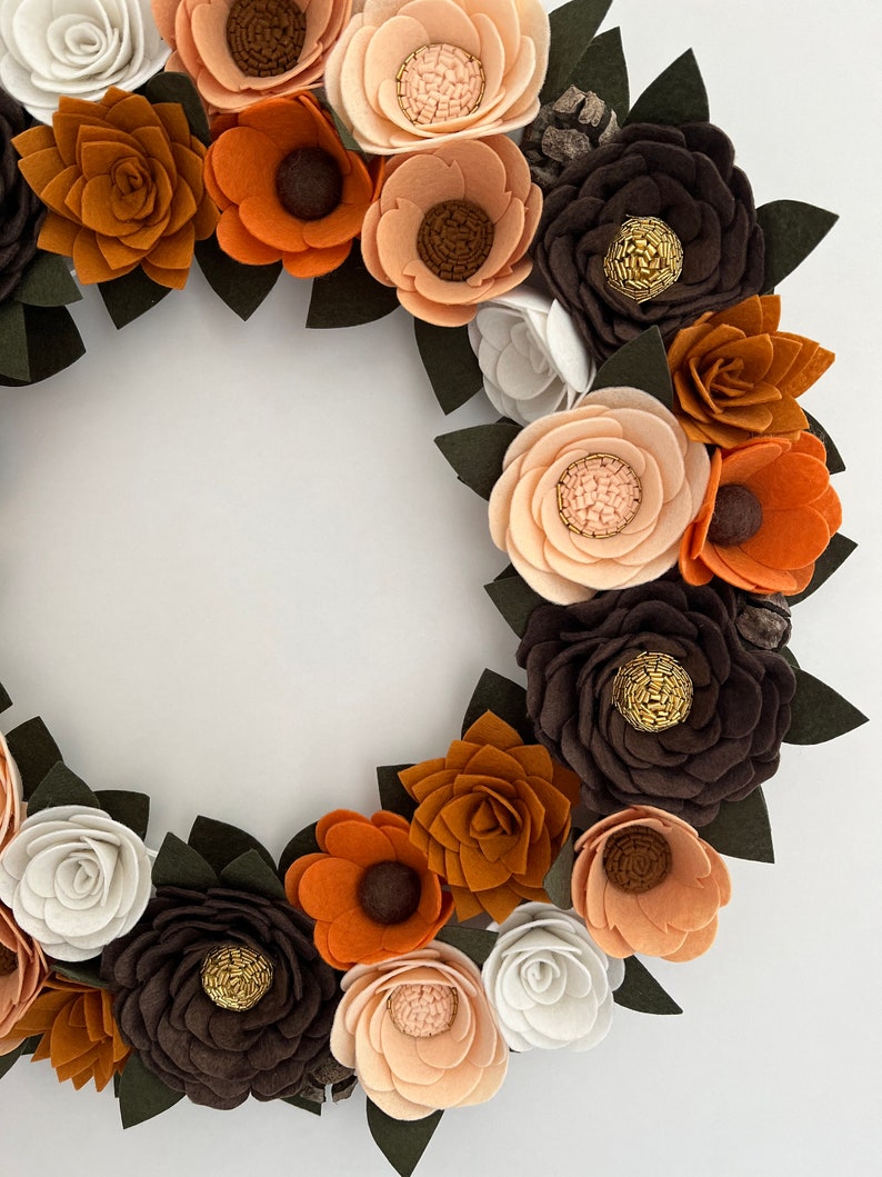 Puede incluir: Una corona de flores de fieltro hecha a mano con flores en tonos marr&oacute;n, naranja y crema, acentuada con hojas de color verde oscuro. La corona circular es ideal para la decoraci&oacute;n de temporada.
