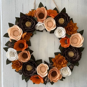 Puede incluir: Una corona circular decorativa con flores de fieltro en tonos oto&ntilde;ales: marr&oacute;n, naranja y crema, con hojas verdes y pi&ntilde;as. Las flores tienen centros detallados, y la corona se cuelga de un lazo de cuerda natural, adecuada para la decoraci&oacute;n estacional.