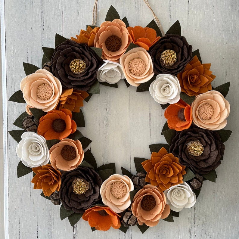 Puede incluir: Una corona circular decorativa con flores de fieltro en tonos oto&ntilde;ales: marr&oacute;n, naranja y crema, con hojas verdes y pi&ntilde;as. Las flores tienen centros detallados, y la corona se cuelga de un lazo de cuerda natural, adecuada para la decoraci&oacute;n estacional.