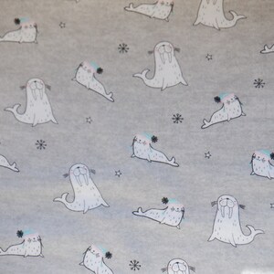 May include: A light gray fabric with a repeating pattern of cartoon walruses wearing hats. The walruses are white with black details and pink cheeks. Small snowflakes and stars are scattered across the background.