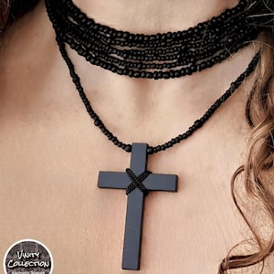 May include: Black beaded necklace with a cross pendant. The necklace features multiple strands of small beads at the choker level, with a single strand extending to a cross pendant. The cross is a solid, dark color and is tied with a black cord.