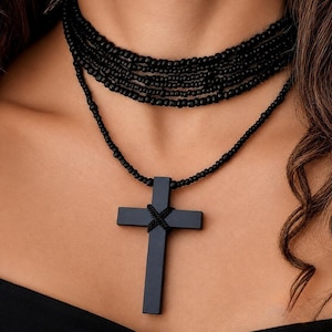 May include: A multi-layered black beaded choker necklace featuring a long strand that suspends a solid black cross pendant. The cross is accented with a small black cord wrapped in an X-shape at the center where the horizontal and vertical bars meet.