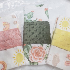May include: Three white fabric burp cloths with different patterns. The first cloth has a pink fuzzy trim and a rainbow and cloud pattern. The second cloth has a green fuzzy trim and a cactus and flower pattern. The third cloth has a yellow fuzzy trim and a sun and cloud pattern.