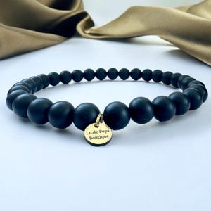 Può includere: Una collana di perline nere con un ciondolo color oro. Il ciondolo è rotondo e reca l'incisione "Little Pups Boutique". Le perline sono opache e di dimensioni variabili, creando un accessorio elegante.