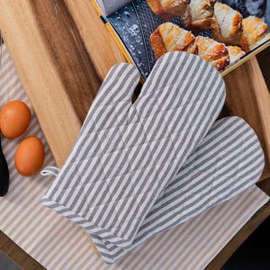 May include: A pair of gray and white striped oven mitts with quilted stitching. The mitts are lying on a striped beige and brown surface.