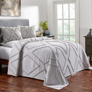 May include: A light grey bedspread and pillow set with geometric patterns. The bedding features a tufted design with a textured appearance. The bedroom setting includes a grey headboard, bedside table, and wooden flooring.
