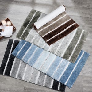May include: A collection of striped bath mats in various colors, including gray, brown, and blue. The mats are arranged on a light-colored wooden surface, with one rolled up. The mats have a soft, textured appearance.