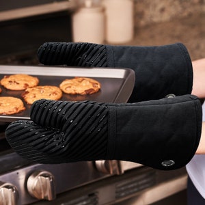 May include: A pair of black oven gloves with a textured, heat-resistant silicone grip. The gloves have a black cotton exterior and a black silicone interior. The gloves are being used to lift a baking sheet with cookies on it.