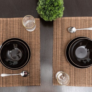 May include: Two place settings with black bowls, brown woven placemats, silver spoons, and clear glasses on a dark wood table.