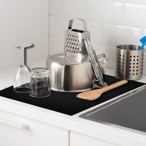 May include: A black silicone kitchen mat with a stainless steel pot, a glass measuring cup, a glass wine glass, a wooden spoon, and a grater on top.