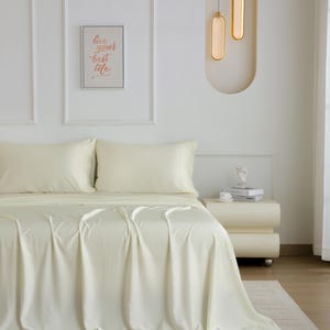 May include: A white bed with a white duvet cover and two white pillowcases. The bed is in a bedroom with white walls and a white bedside table. The bedside table has a book and a small decorative object on it. There are two gold pendant lights hanging from the ceiling.