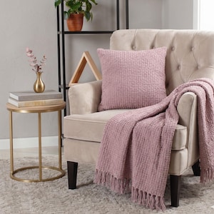 May include: A cozy living room scene featuring a blush-colored textured throw blanket with fringe, a matching square pillow, and a tufted armchair. A small gold side table with books and a vase adds a touch of elegance. A black metal shelf holds a potted plant.