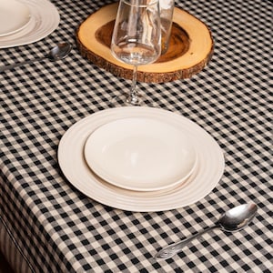 May include: A black and white checked tablecloth with a white plate, a smaller white plate, and a silver spoon. A glass of water and a wooden coaster with dried flowers are also on the table.