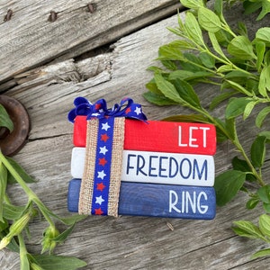 May include: A stack of three wooden blocks painted red, white, and blue with the words "LET FREEDOM RING" in white lettering. The blocks are tied together with a red, white, and blue ribbon with white stars.