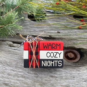 May include: Three wooden blocks stacked on top of each other, tied with twine. The blocks are painted red, white, and black with the words "WARM", "COZY", and "NIGHTS" written in black.