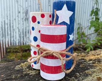 May include: Three wooden firecracker decorations. One is blue with white stars, another is white with red and blue polka dots, and the third is red and white striped. Each has a jute twine fuse and is tied together with more twine. They are displayed on a weathered wooden surface.