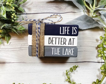 May include: Three wooden blocks stacked on top of each other. The top block is navy blue with the text "LIFE IS" in white. The middle block is white with the text "BETTER AT" in black. The bottom block is gray with the text "THE LAKE" in white. The blocks are tied together with twine.