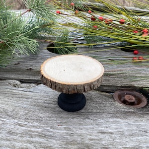 May include: A small wooden cake stand with a natural wood top and a black painted base. The stand is sitting on a weathered wooden surface.