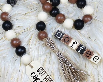 May include: A wooden bead garland with brown, black, and white beads. The garland has a brown and white tassel and a wooden sign that says "But First Coffee". There is also a wooden block sign that says "Coffee".
