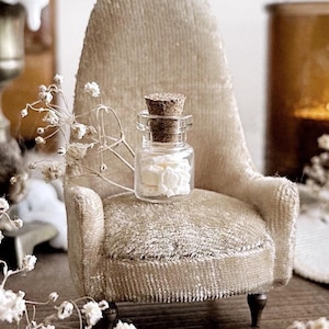 May include: A small, white, velvet armchair with a glass bottle filled with white objects on the seat. The bottle has a cork stopper and is sitting on a white, velvet cushion. The chair is in a room with white walls and a wooden floor.