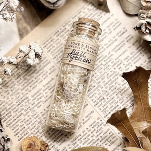 May include: A glass jar filled with a white, textured material, labeled "Ball Python" and "BONE & BLOOM CURIOSITY MARKET". The jar is sitting on a book with a brown background.