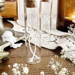 May include: Two clear glass vials with cork stoppers, each containing a white, decorative, intertwined design. The vials sit on a wooden surface, with a lit candle in a brown glass holder and dried flowers in the background. A vintage aesthetic.