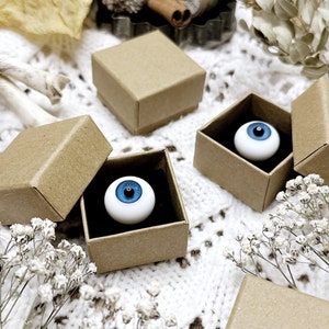 May include: Three small brown cardboard boxes with lids open, each containing a white eyeball with a blue iris. The boxes are on a white crocheted surface.