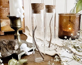 May include: Two clear glass bottles with cork tops and a single twig inside each. The bottles are sitting on a white lace tablecloth with small white flowers scattered around them.