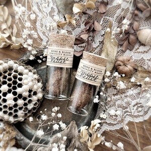 May include: Two small glass vials with cork stoppers, each labeled "Bone & Bloom Curiosity Market Moose Pellet." The vials contain a brown pellet and are set on a decorative tray with dried flowers and lace.