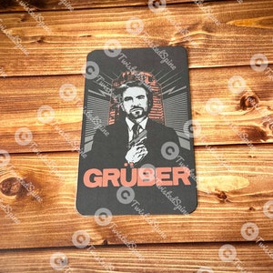 May include: A black and white illustration of a man with a gun, with the text "GRUBER" in red letters.