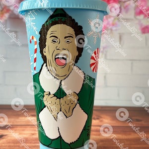 May include: A blue reusable cup with a white lid and a green and white image of Buddy the Elf from the film Elf. The image shows Buddy with his arms crossed and a big smile on his face. The cup has a white snowflake design and a red and white candy cane design.
