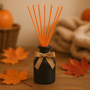 May include: A black aromatherapy diffuser bottle with orange reeds and a gold ribbon. The bottle sits on a wooden surface with orange maple leaves. A pumpkin and a woven basket are in the background.