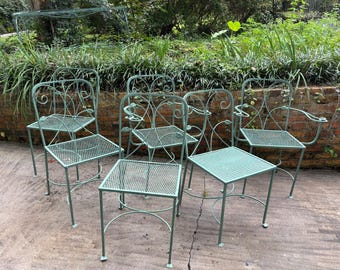 May include: A set of six green metal patio chairs and two matching square tables. The chairs have decorative scrollwork and a mesh seat and back. The tables also have a mesh top. The furniture is outdoors.
