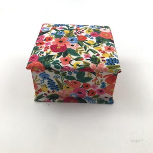 May include: A small, rectangular box covered in a floral fabric. The fabric features a white background with a pattern of colorful flowers in shades of pink, red, blue, yellow, and orange, with green leaves. The box has a simple, clean design.