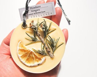 May include: A round, cream-colored air freshener with a sprig of rosemary and a dried lemon slice embedded in the wax. The tag reads "Pleiadians Rosemary & Lemon Scented Gum Air Freshener". A black and silver ribbon is attached.