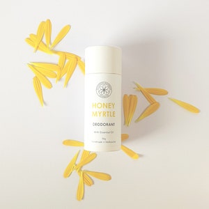May include: White deodorant stick with the text "Honey Myrtle Deodorant With Essential Oil 50g Handmade in Melbourne" on the label. The stick is surrounded by yellow flower petals on a white background.